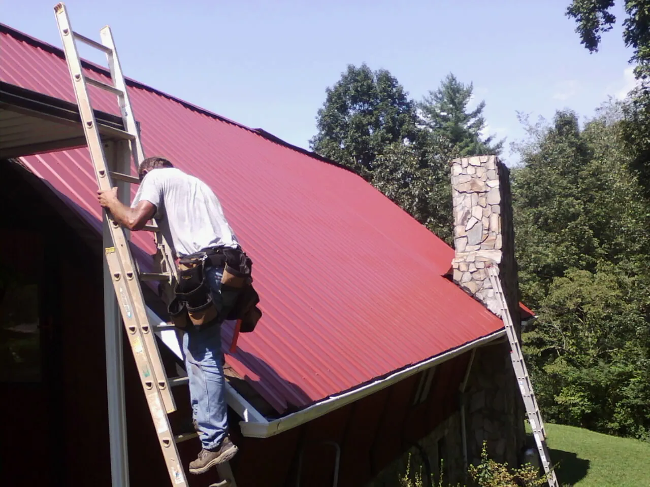 Roofer on a ladder beside a red metal roof and stone chimney — Soffit & Fascia Repair by Roofing Brothers in Manquin
